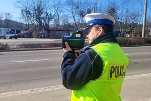 Umundurowany policjant stoi przy jezdni i za pomocą urządzenia trzymanego w rękach mierzy prędkość pojazdów.