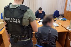 Pomieszczenie służbowe. Policjant w stroju cywilnym i z założoną kamizelką z napisem POLICJA stoi przy zatrzymanym mężczyźnie, który siedzi na krześle. Na przeciwko niego za biurkiem siedzi umundurowany policjant.