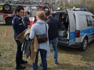 Umundurowana policjantka rozmawia z osobami. W tle oznakowany radiowóz, w którym siedzi dziecko