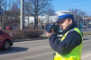 Umundurowany policjant stoi przy jezdni i trzyma w ręku urządzenie do pomiaru prędkości pojazdów