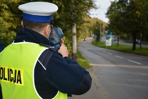 Umundurowany policjant stoi przy jezdni i za pomocą urządzenia trzymanego w rękach mierzy prędkość pojazdu jadącego ulicą