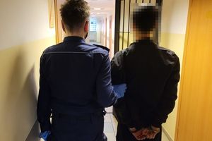 Umundurowana policjantka prowadzi zatrzymanego mężczyznę, który ma założone kajdanki na rękach trzymanych z tyłu