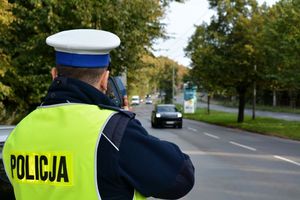 Umundurowany policjant ruchu drogowego stoi przy jezdni i za pomocą urządzenia trzymanego w rękach  mierzy prędkość samochodów jadących w jego stronę.