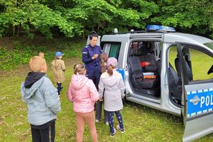 Umundurowana policjantka rozmawia z dziećmi. Za nimi radiowóz i drzewa