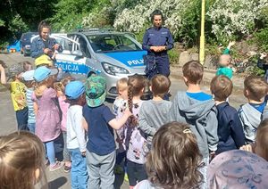 Umundurowana policjantka rozmawia z grupą dzieci i ich opiekunką. Obok stoi oznakowany radiowóz