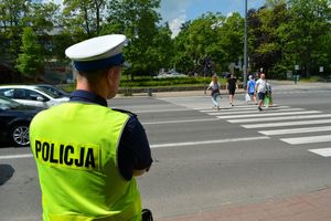 Umundurowany policjant stoi przy skrzyżowaniu i obserwuje osoby idące po przejściu dla pieszych