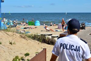 Pora letnia. Umundurowana policjantka obserwuje osoby na plaży. W tle na wodzie widoczne pływające łódki. - zdjęcie archiwalne