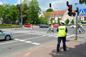 Pora dzienna. Umundurowany policjant stoi przy oznakowanym przejściu dla pieszych i obserwuje osoby idące po przejściu.