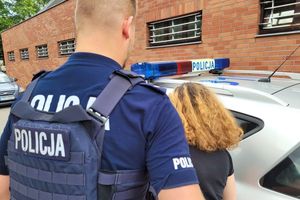 Umundurowany policjant wprowadza do oznakowanego radiowozu zatrzymaną kobietę. W tle ściana budynku Komendy Miejskiej Policji w Sopocie i samochód osobowy