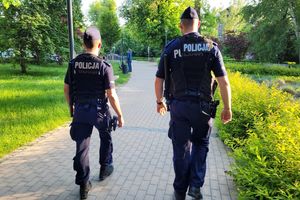 Umundurowani dwaj policjanci idą chodnikiem przez park.