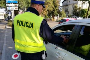 Umundurowany policjant stoi przy samochodzie i za pomocą urządzenia trzymanego w ręce bada trzeźwość osoby kierującej samochodem