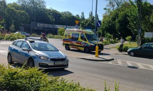 Na zdjęciu fragment ronda i ulicy z przejazdem rowerowym i przejściem dla pieszych. Z lewej strony na chodniku stoi oznakowany radiowóz, a obok niego strażak. Po drugiej stronie na ulicy stoi karetka pogotowia, obok której na chodniku stoją dwie osoby. Obok karetki widoczny fragment samochodu osobowego.