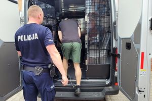 Umundurowany policjant wprowadza do samochodu do pomieszczenia dla osób zatrzymanych mężczyznę, który ma założone kajdanki na nogi i ręce.