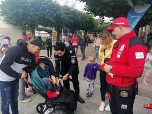 Umundurowana policjantka wręcza dziecku opaskę. Obok stoi ratownik wodny i inni ludzie.