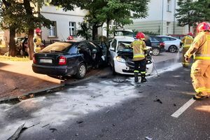 Miejsce zdarzenia drogowego. Na pierwszym planie strażacy pracujący na miejscu zderzenia dwóch samochodów. W tle inne samochody i budynki.