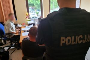 Pomieszczenie służbowe. Umundurowany policjant stoi , a przed nim na krześle siedzi zatrzymany mężczyzna. Za nimi przy biurku siedzi policjant w stroju cywilnym.