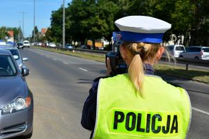 Umundurowana policjantka stoi przy jezdni i mierzy prędkość jadących ulicą pojazdów