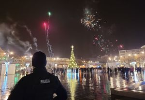 Pora nocna. Umundurowany policjant obserwuje osoby stojące na placu. W tle wystrzelone w górę fajerwerki