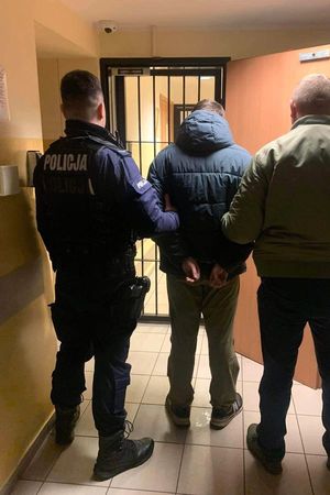 Umundurowany policjant i policjant w stroju cywilnym stoją z zatrzymanym mężczyzną, który ma założone kajdanki na ręce trzymane z tyłu przed pomieszczeniem dla osób zatrzymanych