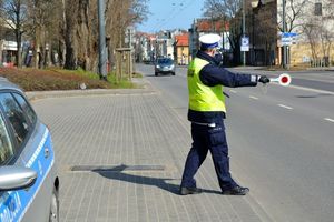 Umundurowany policjant stoi przy ulicy i trzymając w ręku tarczę do zatrzymywania pojazdów zatrzymuje jadący ulicą pojazd. Z lewej strony widoczny fragment oznakowanego radiowozu. Zdjęcie archiwalne