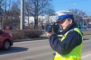 Pora zimowa. Umundurowany policjant stoi przy jezdni i trzyma w ręku urządzenie do pomiaru prędkości jadących pojazdów.