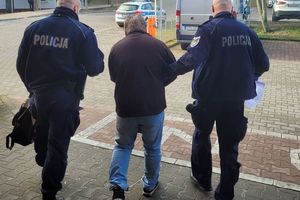 Dwaj umundurowani policjanci prowadzą zatrzymanego mężczyznę. Za nimi w tle parkingi i zaparkowane na nich pojazdy