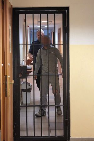 Pomieszczenie dla osób zatrzymanych. Umundurowany policjant prowadzi korytarzem zatrzymanego mężczyznę.