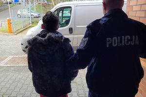 Umundurowany policjant prowadzi zatrzymanego policjanta. Za nimi w tle samochody i parking