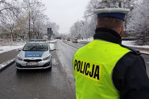 Pora zimowa. Umundurowany policjant ruchu drogowego stoi przy ulicy i obserwuje jadące pojazdy. Za nim w zatoczce oznakowany radiowóz