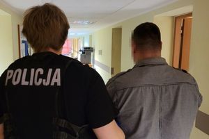 Policjant w stroju cywilnym i z założoną kamizelką z napisem POLICJA prowadzi zatrzymanego mężczyznę