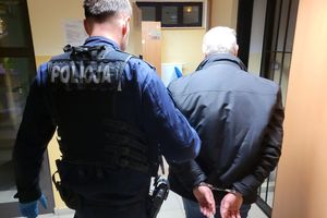 Umundurowany policjant prowadzi zatrzymanego mężczyznę, który ma założone kajdanki na ręce trzymane z tyłu