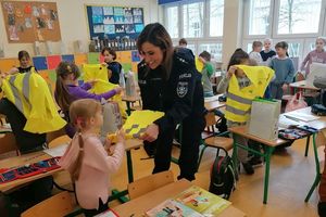 Wnętrze szkolnej klasy. Umundurowana policjantka wręcza dzieciom siedzącym i stojącym przy ławkach kamizelki odblaskowe.