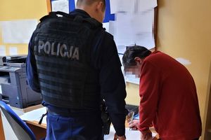 Pomieszczenie służbowe. Umundurowany policjant stoi przy biurku. Obok niego stoi mężczyzna w stroju cywilnym i pisze na dokumencie, który leży na biurku.