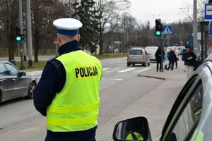 Umundurowany olicjant stoi przy jezdni, obok radiowozu. Za nim widoczna ulica, po której jada samochody oraz przejście dla pieszych, przy którym stoją piesi.