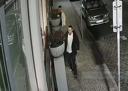 Pora nocna. Dwóch mężczyzn stoi na chodniku przy budynku, z któych jeden schowany jest częściowo za donicę stojącą na chodniku. Obok chodnika na jezdni stoją zaparkowane samochody.