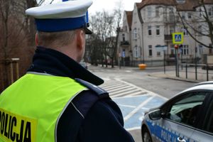 Umundurowany policjant stoi obok rdiowozu i obserwuje skrzyżowanie ulic, gdzie są też przejścia dla pieszych