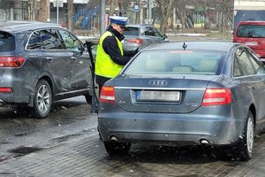 Umundurowany policjant stoi przy samochodzie i kontroluje kierującego samochodem. Za nimi ulica i jadące po niej inne samochody.