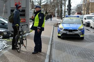 Umundurowanny policjant rozmawia z rowerzystą. Oboje stoją na chodniku. Za nimi widoczny oznakowany radiowóz i z prawej strony ulica, po której jadą samochody.