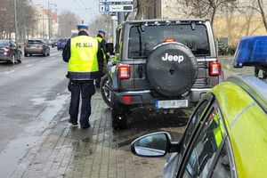 Umundurowani policjanci stoją przy samochodach i kontrolują kierujących. Z lewej strony widoczna ulica i jadące po niej inne samochody. Przed policjantami i kontrolowanymi pojazdami z prawej strony widoczny fragment oznakowanego radiowozu.