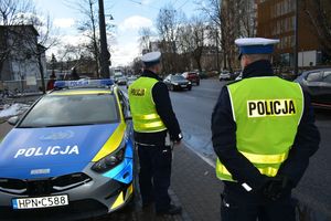 Pora dzienna. Dwóch umundurowanych policjantów stoi przy jezdni i obserwuje jadące ulicą samochody. Obok policjantów w zatoczce stoi oznakowany radiowóz.