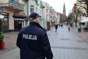 Umundurowany policjant idzie w górę ulicy. Z naprzeciwka idą inne osoby, a po bokach i w tle widoczne są budynki z lokalami usługowymi oraz wieża kościoła