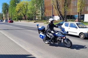 Umundurowany policjant na oznakowanym motocyklu jedzie ulicą. Obok i za nim widoczne inne samochody jadące ulicą.