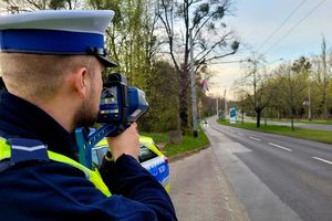 Umundurowany policjant za pomocą urzędzenia trzymanego w rękach mierzy prędkość jadącego ulica samochodu.