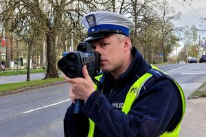 Umundurowany policjant stoi przy ulicy i za pomocą urzędzenia trzymanego w rękach mierzy prędkość.