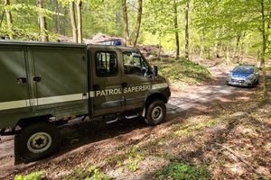 Teren leśny z widoczną drogą, na której z lewej strony stoi zaparkowany pojazd wojskowy z napisem PATROL SAPERSKI i w tle po prawej stronie stoi zaparkowany oznakowany raiowóz.