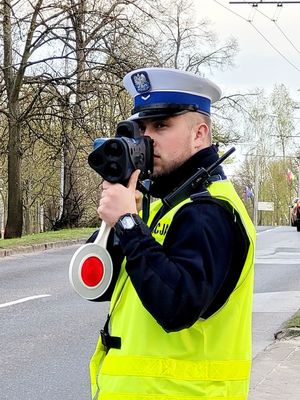 Umundurowany policjant stoi przy jezdni i trzyma w rękach urządzenie do pomiaru prędkości oraz przedmiot w kształcie lizaka będący tarczą do zatrzymywania pojazdów