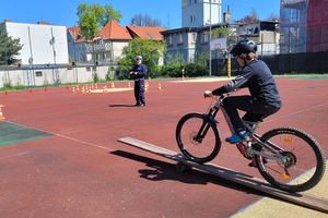 Na zdjęciu boisko szkolne. Młoda osoba na rowerze jedzie to torze sprawnościowym, a jego przejazd obserwuje stojący obok umundurowany policjant.