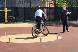 Na zdjęciu boisko szkolne. Młoda osoba na rowerze jedzie to torze sprawnościowym, a jego przejazd obserwuje stojący obok umundurowany policjant.