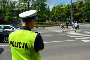 Umundurowany policjant obserwuje uczestników ruchu. Za nim w tle przejście dla pieszych, po którym idą osoby oraz stojące przy skrzyżowaniu osoby