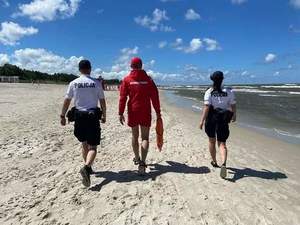 Na zdjęciu plaża, a na niej umundurowani policjanci i ratownik wodny, którzy idą przy wodzie wzdłuż plaży